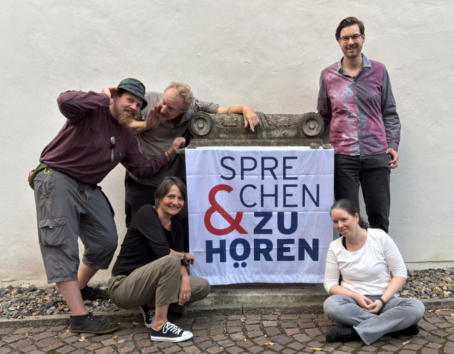 fünf Personen sitzen und stehen in lockerer und lustiger Atmosphäre um das Logo auf einem Banner. Eine Person flüstert der anderen etwas zu.